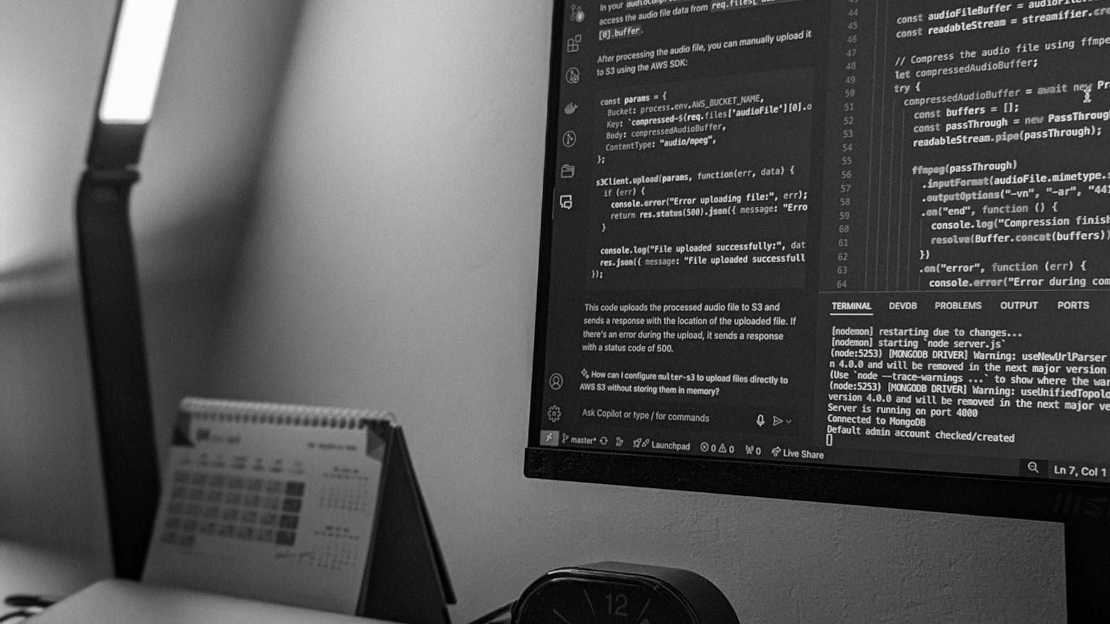 Espacio de trabajo minimalista en blanco y negro con un monitor mostrando líneas de código, un portátil cerrado y una lámpara de escritorio